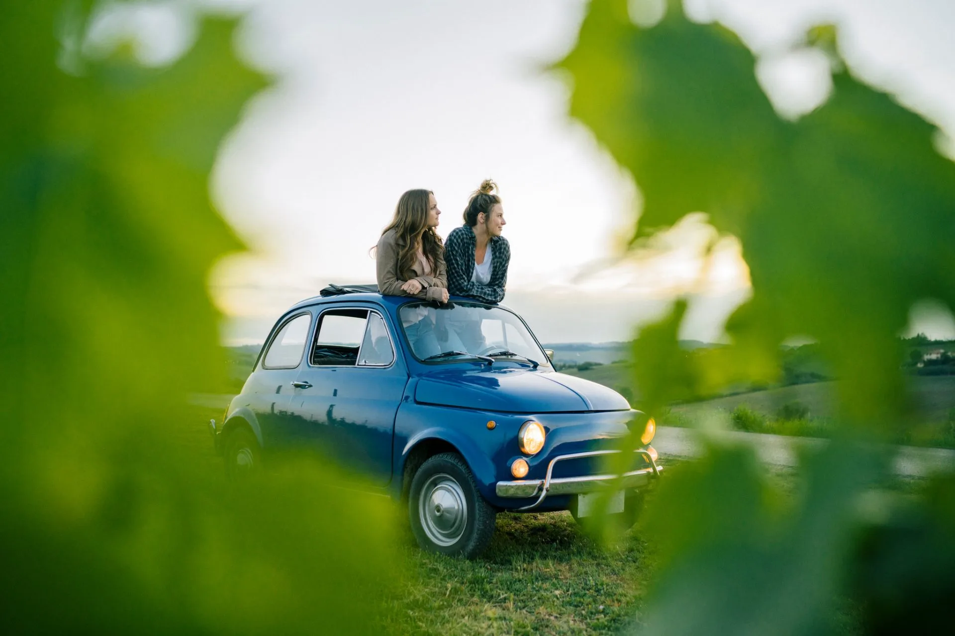 Due amiche con un'auto blu in un paesaggio collinare